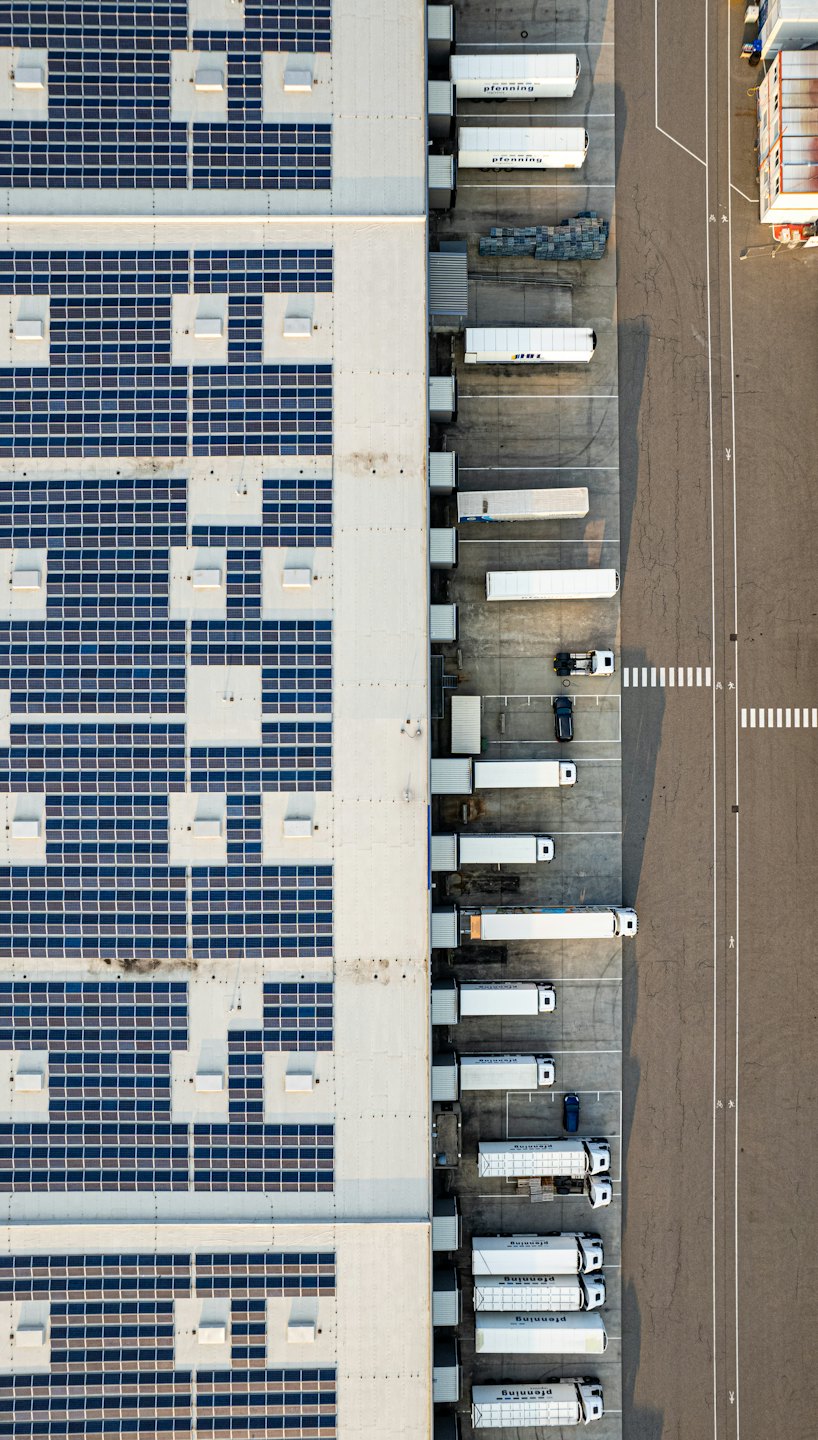 Solar panels cover the flat roof of an industrial facility near semi-trucks at a loading dock.